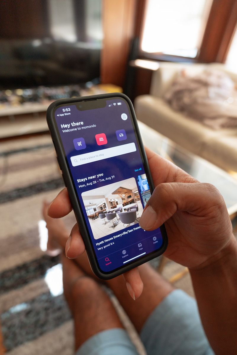 A POV-shot of a man's hand holding up an iPhone showing hotel search results times in the Momondo app, with a soft-focus living room in the background.