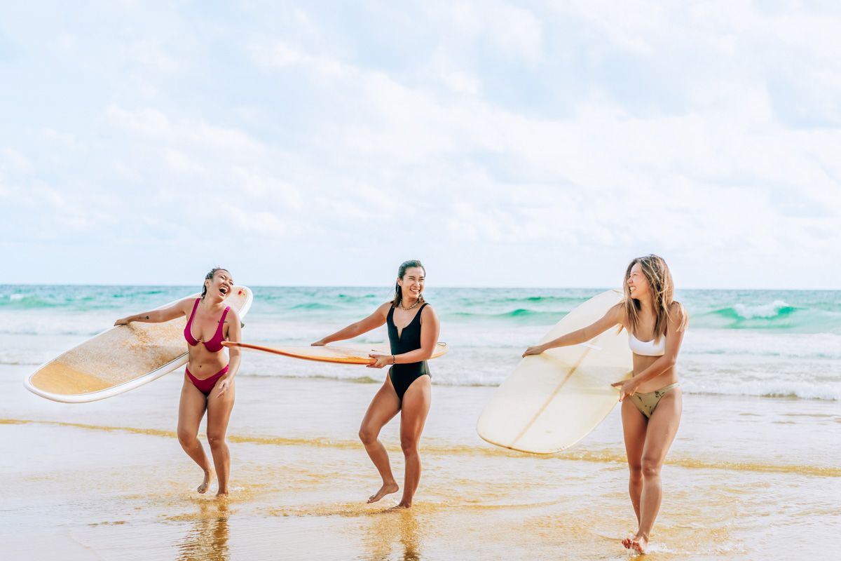 Three female surfers, two in bikinis and one in a one-piece, laugh together as they walk out of the waves back onto the beach.