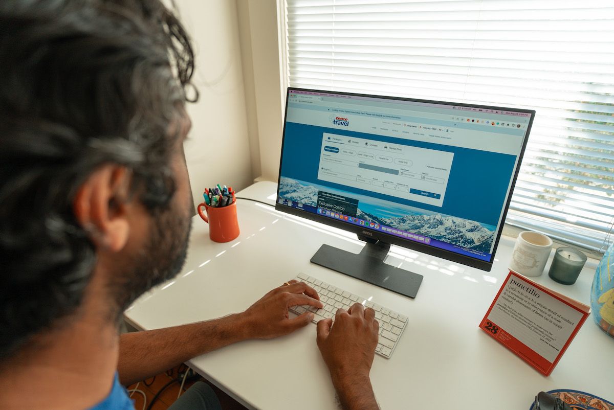 A view over an man's shoulder as he types on a keyboard in front of a monitor sitting on a white desk displaying the Costco Travel website.
