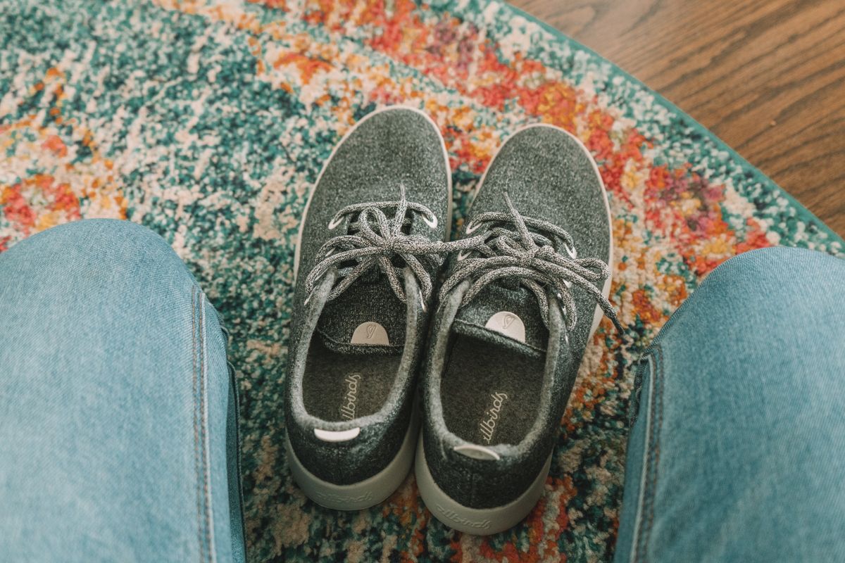Grey Allbirds Wool Runners sitting on a colorful carpet, framed my the photographer's legs.