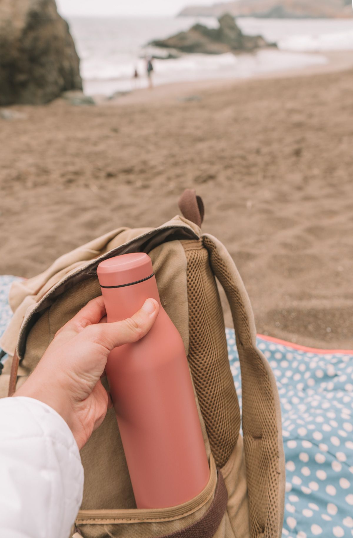 A hand holds a pink water bottle against a light brown backpack with an overcast beach in soft-focus in the background.