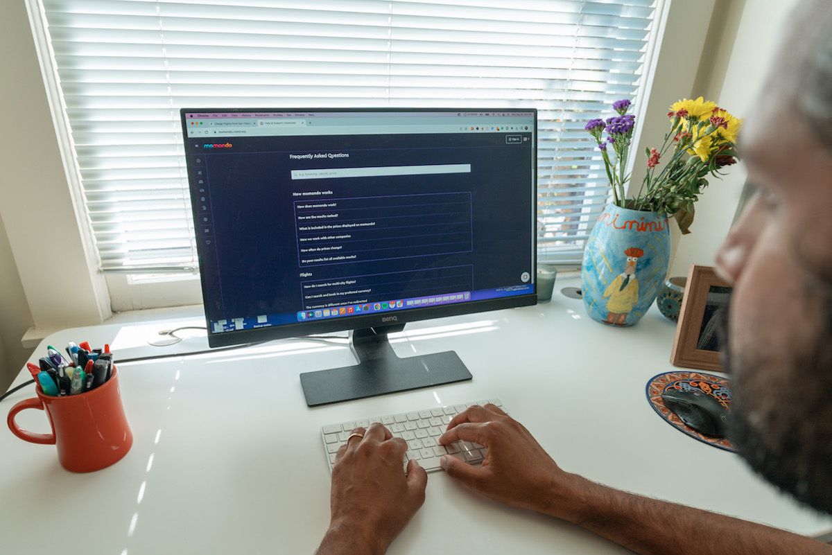 A view looking over the shoulder of a man sitting at a white desk, looking at a computer monitor displaying the FAQs page on the Momondo website.