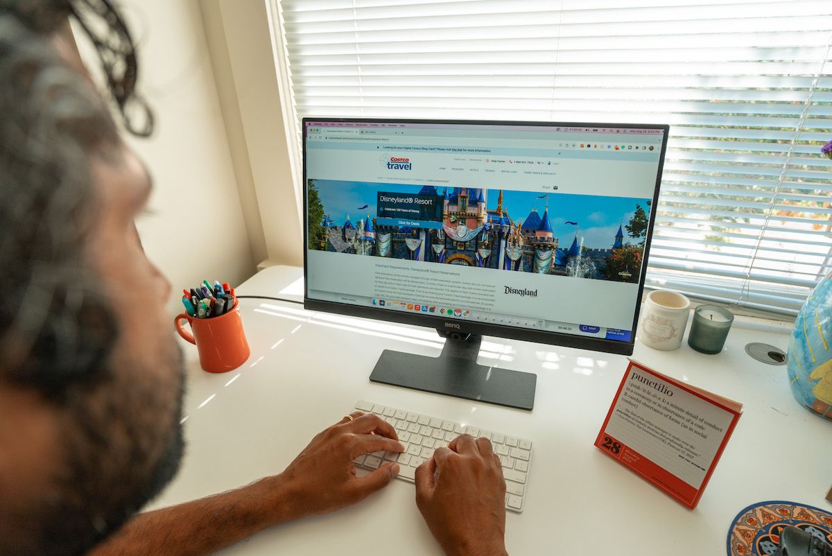 A view over a man's shoulder as he types on a keyboard in front of a monitor displaying the Costco Travel website sitting on a white desk while testing it out during this Costco Travel review.