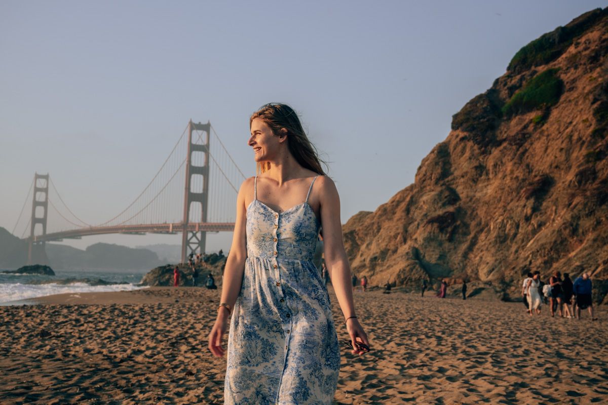 A woman in a blue and white sleeveless sundress standing on the beach and looking over her right shoulder with the Golden Gate Bridge behind her, showcasing some items from our beach packing list.