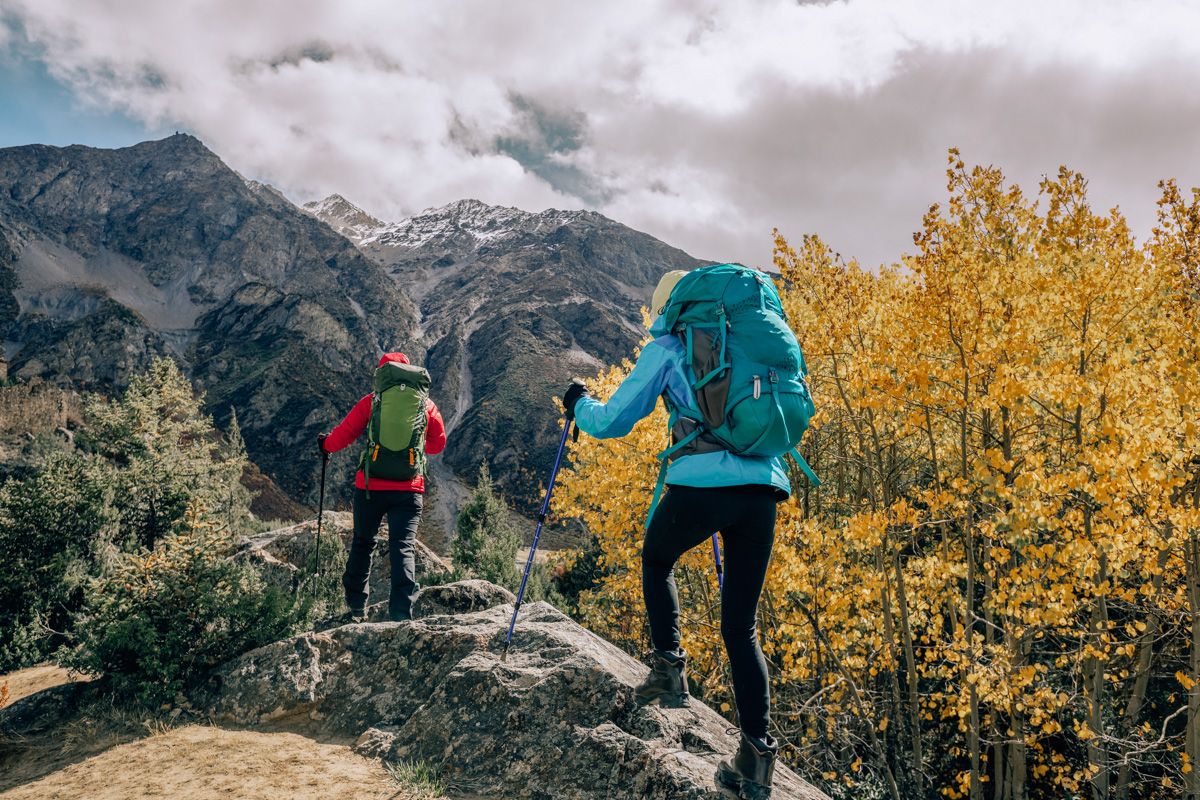 Two hikers wearing two of the best Arc'teryx jackets seen from behind wearing brightly-colored windbreakers and backpacks as they hike over a large boulder, with a mountain and yellow foliage beyond.