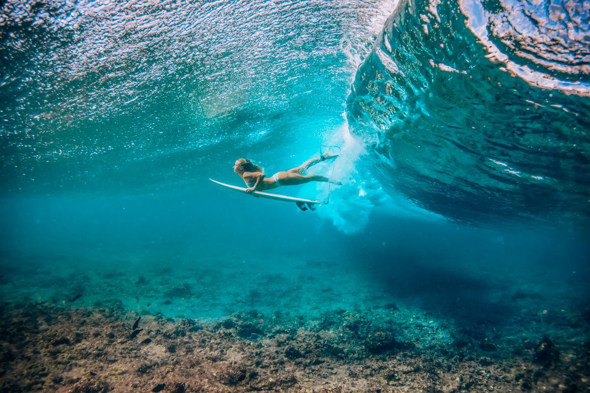 A female surfer wearing one of the best surf bikinis, photographed underwater, swimming with her surfboard in hand.