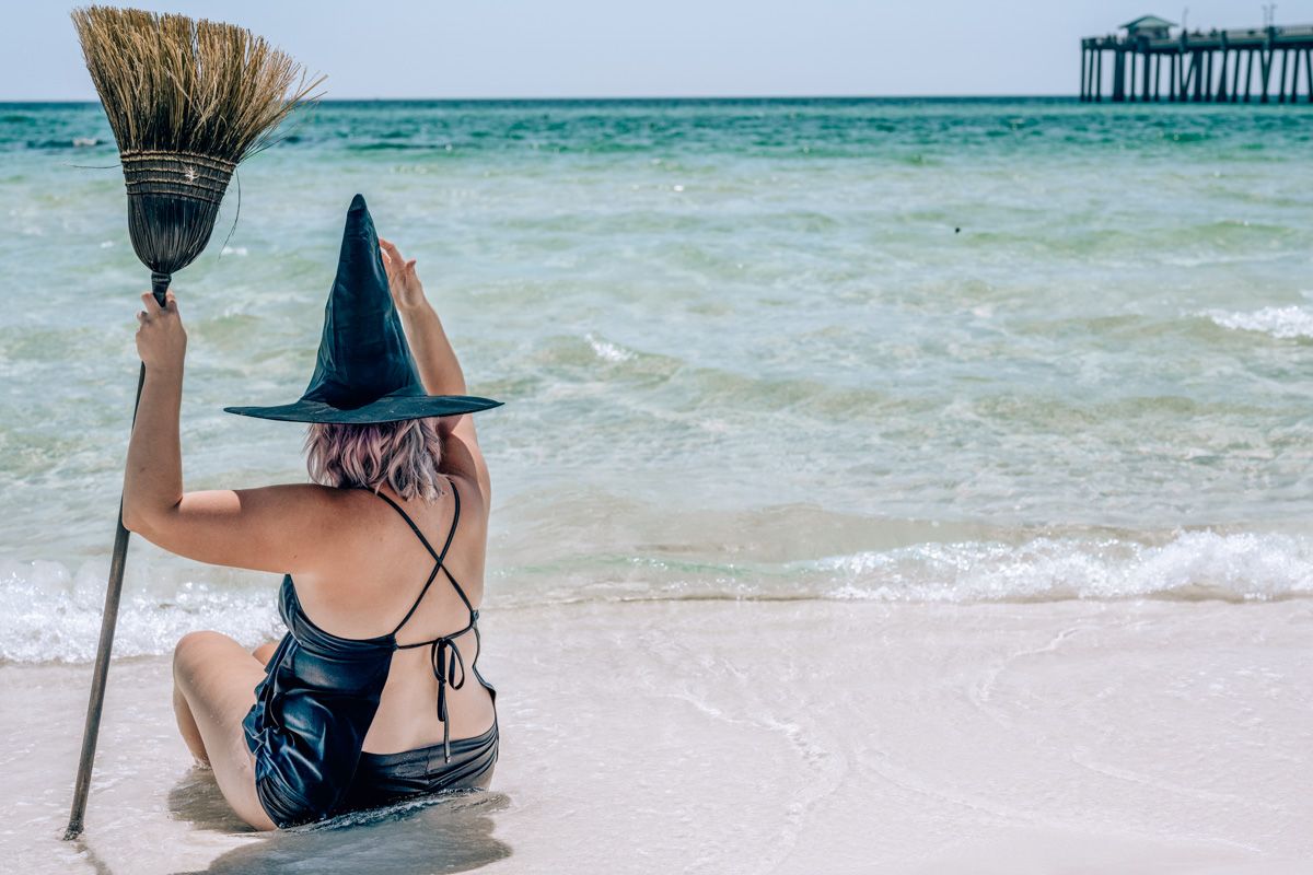 A woman in a black bathing suit and a witch's hat seen from behind as she holds a broom and sits on a sunny California beach.