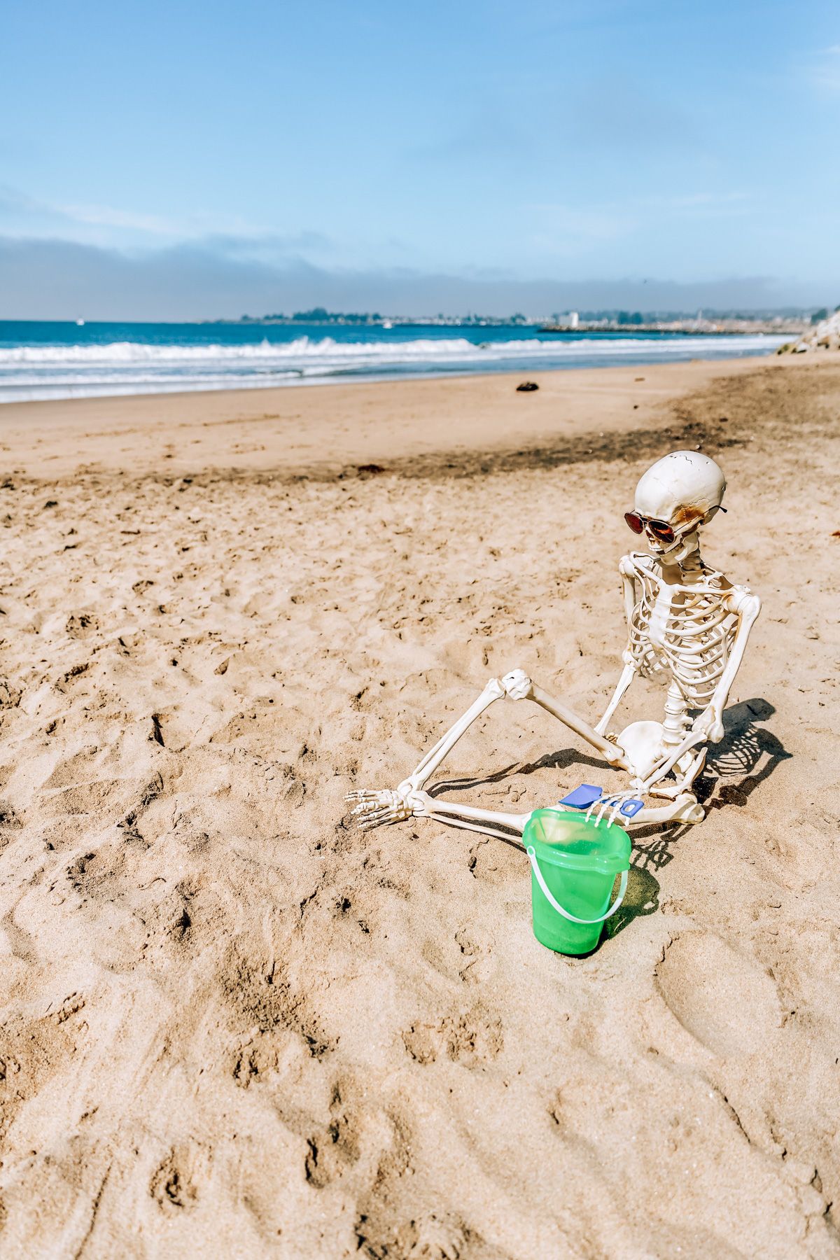 Halloween Events in San Diego: a skeleton wearing sunglasses posed sitting on a a Southern California beach, with a bucket and shovel in hand.