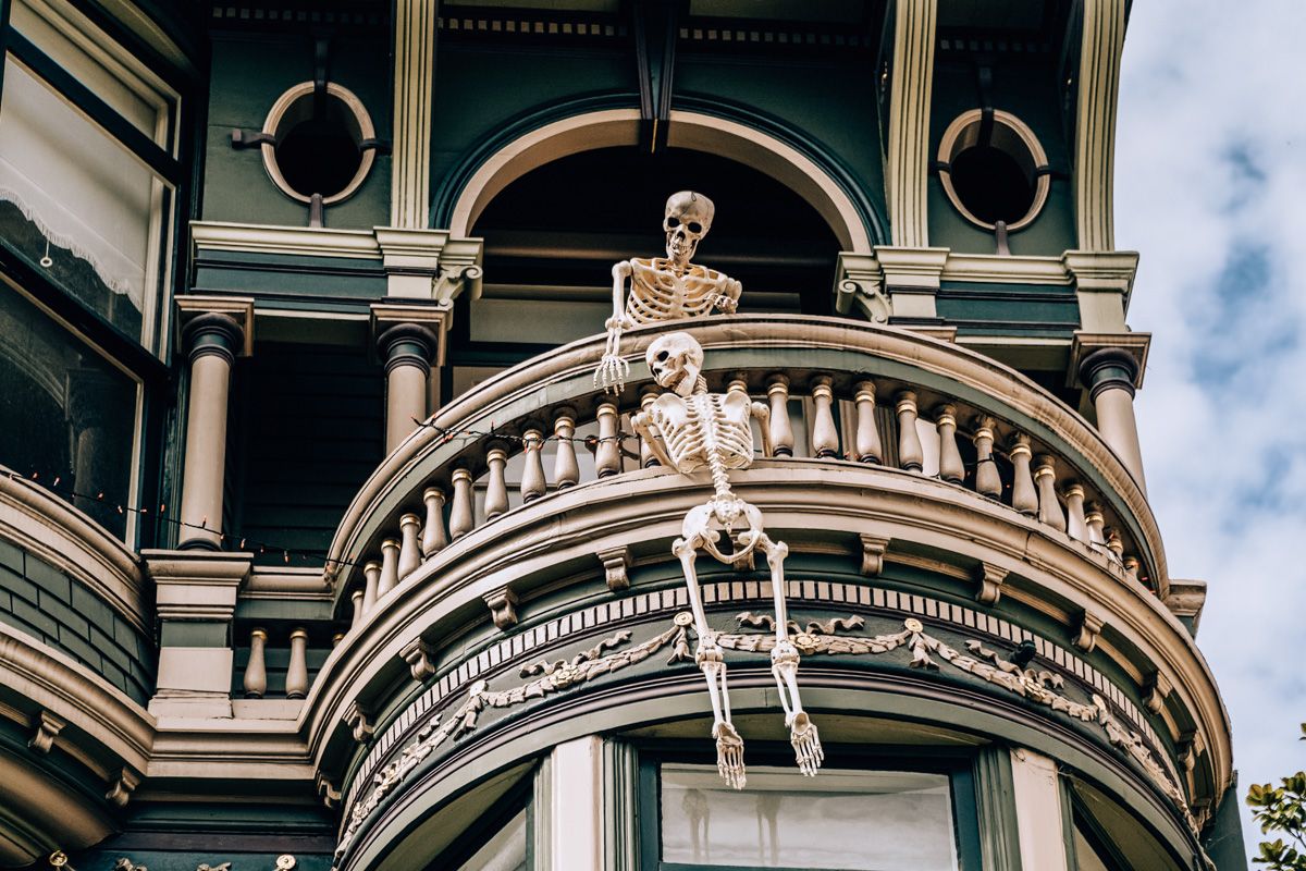 A pair of skeleton halloween decorations posed dangling over the ornate balcony of a Victorian mansion.