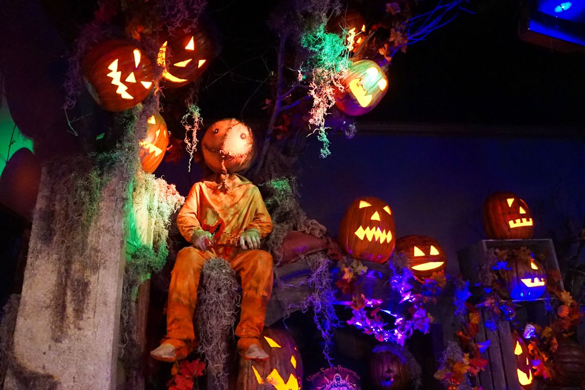 A figure dressed in an orange jumpsuit with a pumpkin head sits amongst illuminated jack-o-lanterns, moss, and colored lights.
