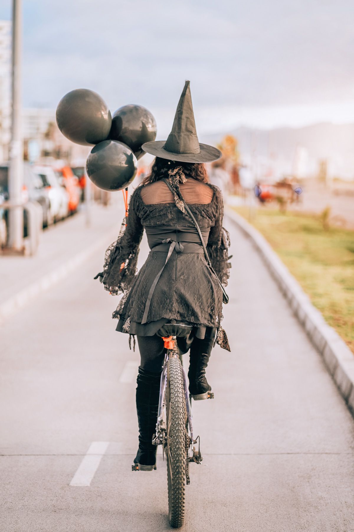 A bicycling woman seen from behind, dressed as a witch for Halloween in Los Angeles, and carrying a bundle of three black balloons.