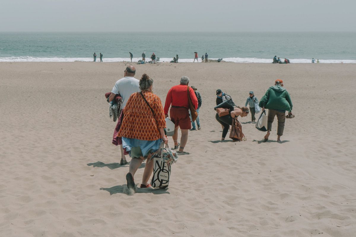 A group of five people seen from behind, walking on the beach towards the water, overloaded with bags and gear. 