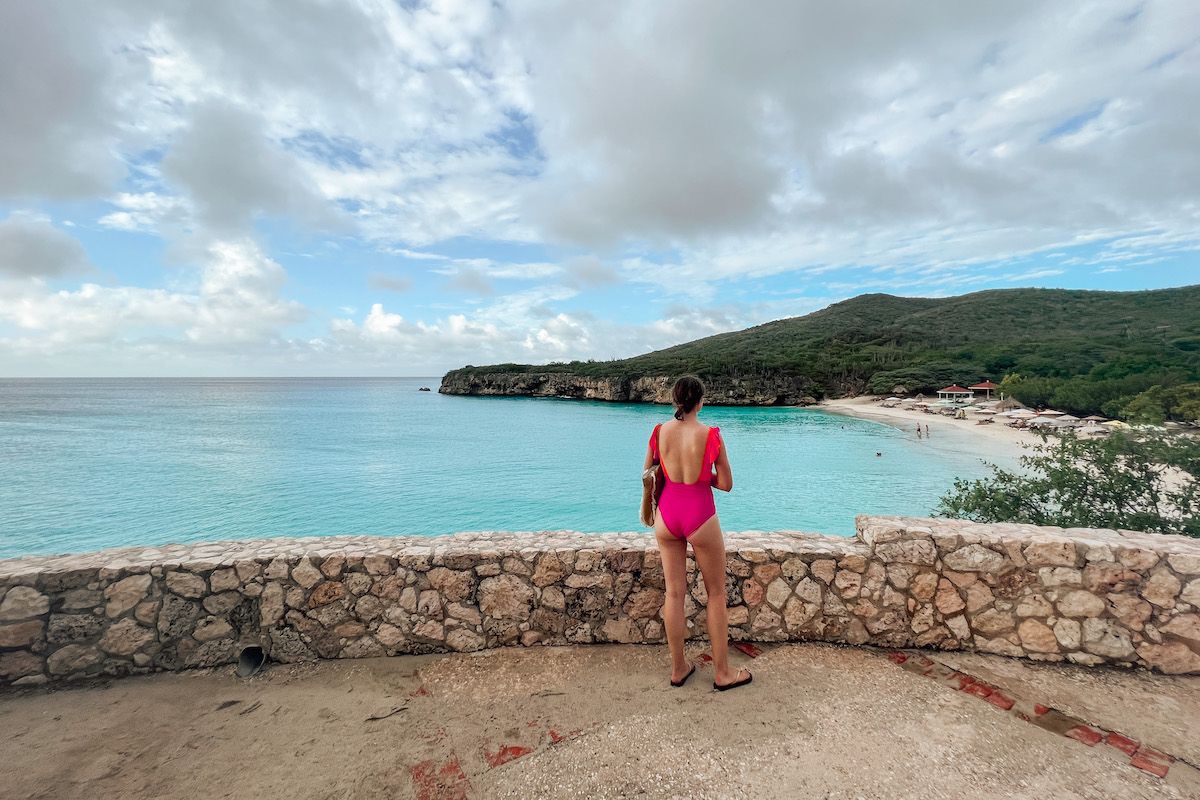 A woman in a magenta one-piece swimsuit seen from behind looking out over a low stone wall at the turquoise waters of a tropical beach. 