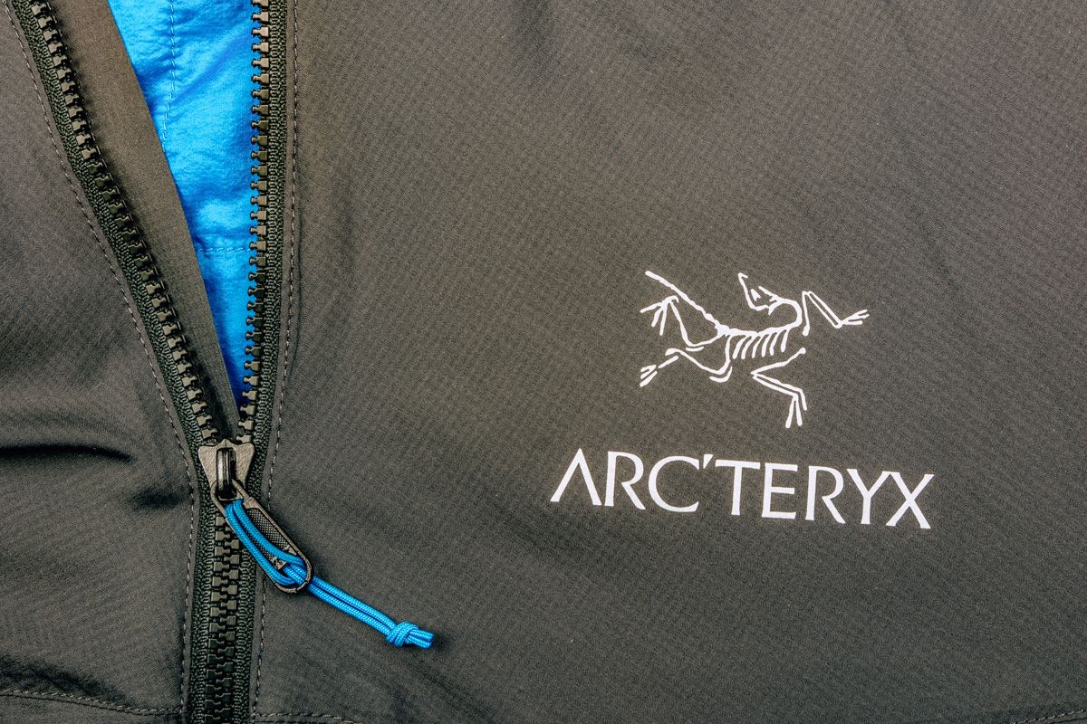 A close-up of an Arc'teryx logo on the front of a black jacket next to the zipper.
