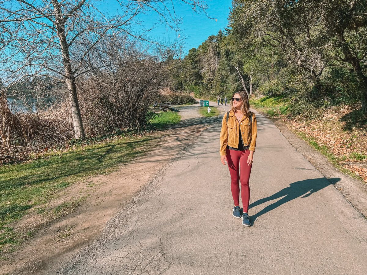 A woman dressed in merlot leggings and a tan corduroy jacket walking a paved nature path along the river in California, wearing Allbirds Dashers shoes.