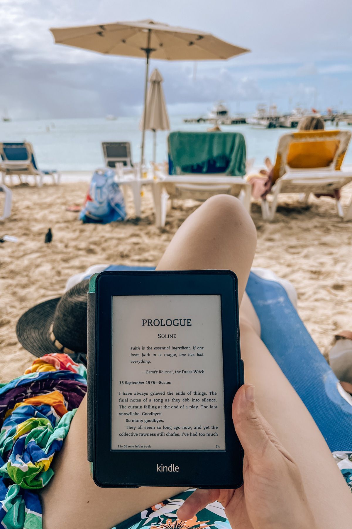 A POV shot of a hand holding a kindle displaying the first page of a book, a pair of legs lounging on a beach chair and a beach in soft-focus visible beyond.