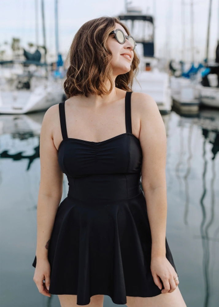 Product image for the Jessica Rey Emma Swim Dress in black, modeled by a woman with shoulder-length brown hair, standing in front of a harbor.