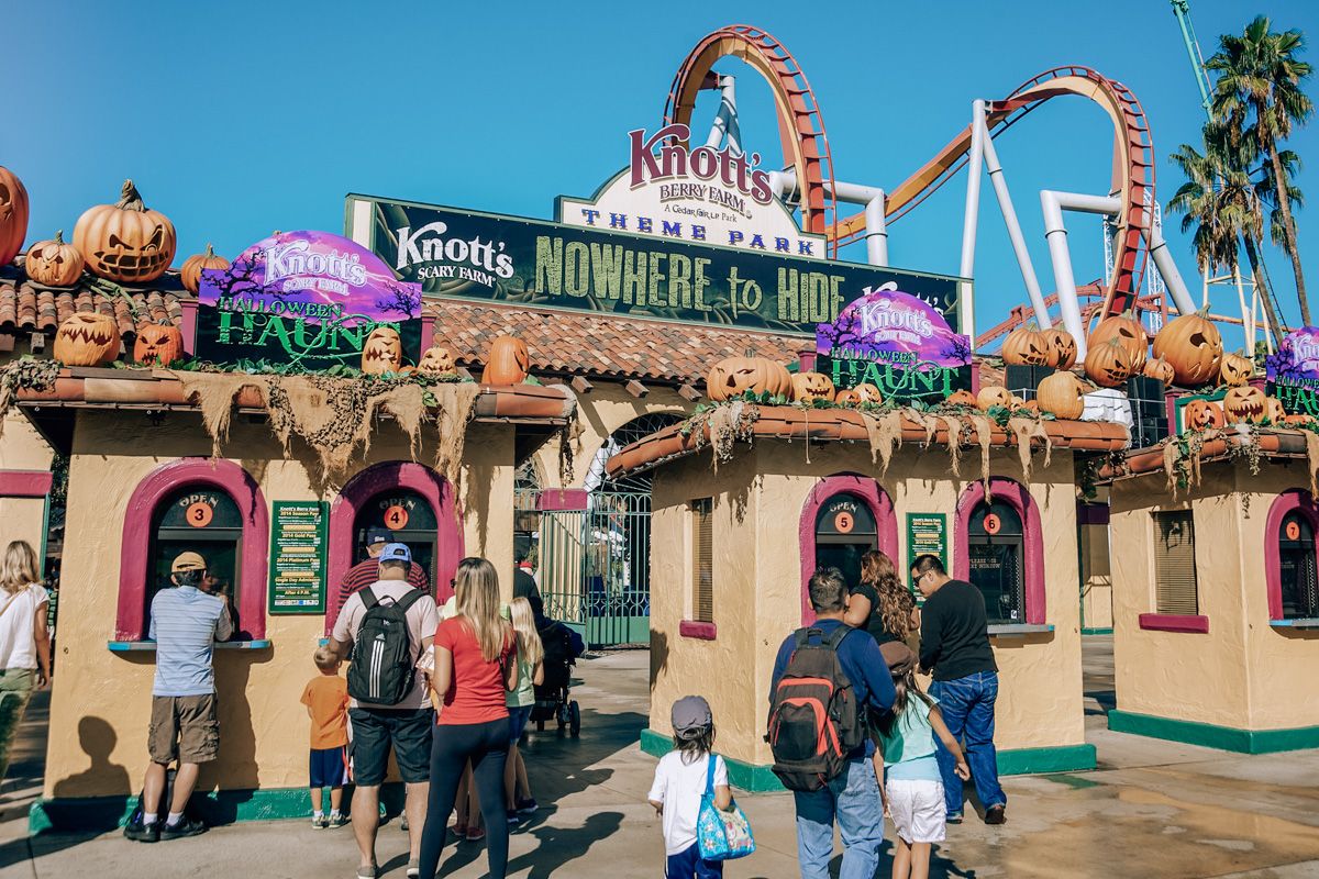 People waiting at the ticket counter at Knott's Scary Farm, with a roller coaster against a clear blue sky in the background.