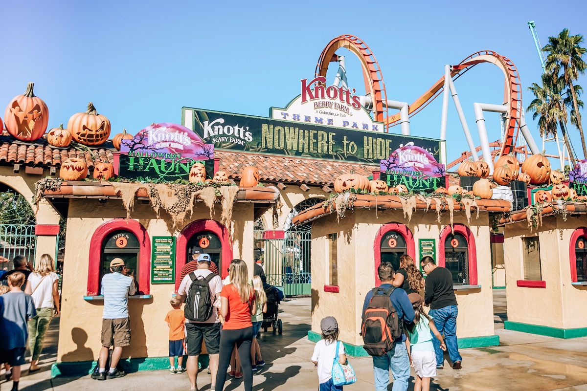 People waiting at the ticket counter for Knotts Scary Farm at Buena Park, with a roller coaster visible against a clear blue sky in the background.