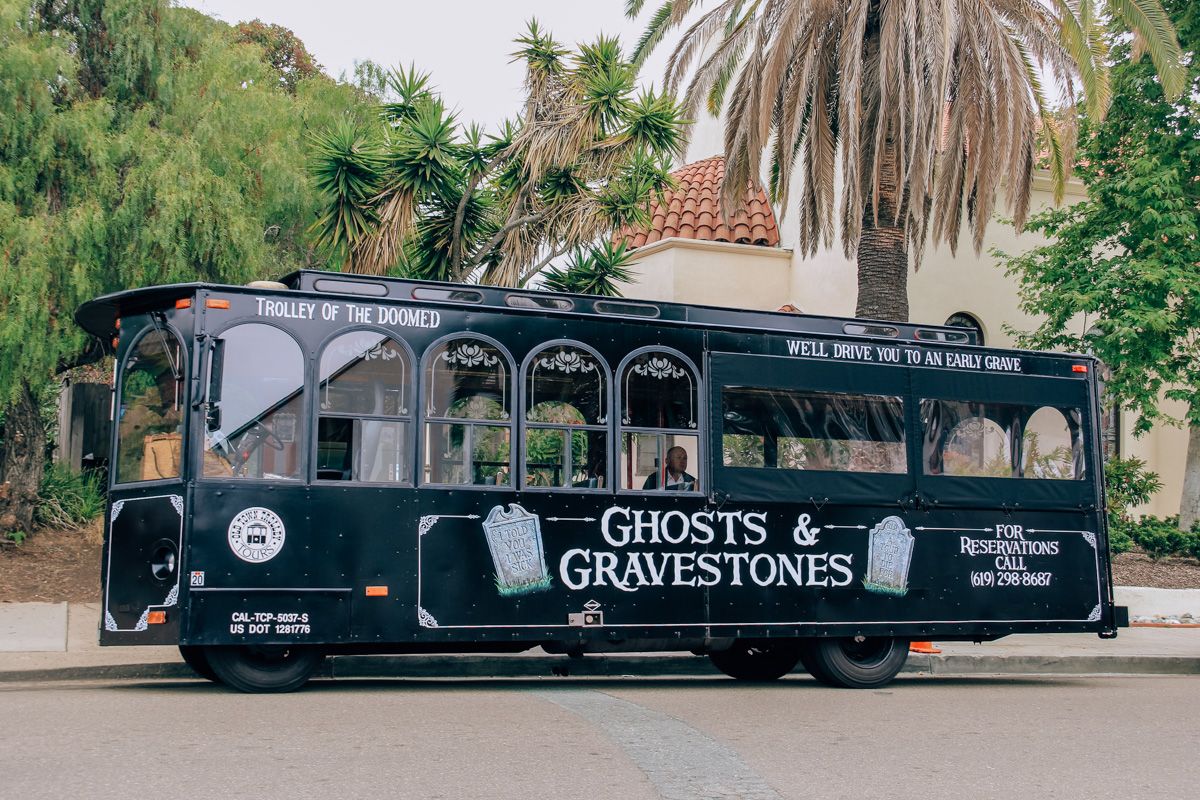 The black bus for the Old Town Trolley Ghost and Gravestone Tours seen parked by the curb of a street, with palm trees and a Spanish-style tile roof visible behind it.