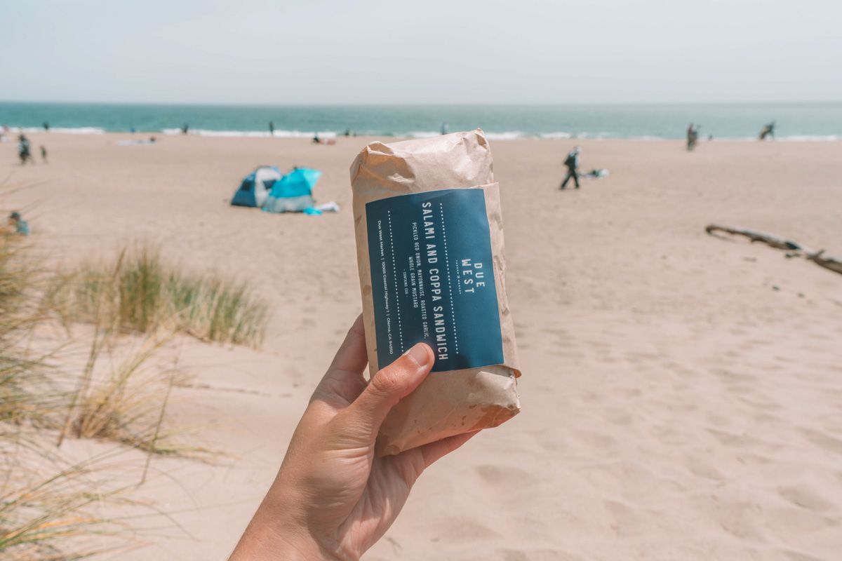 A hand holds up a sandwich wrapped in brown paper labeled, "salami and coppa sandwich" with a beach in the background.
