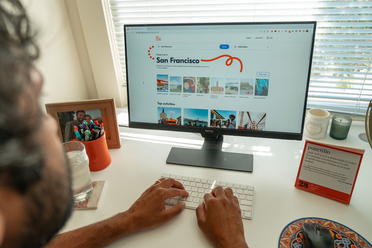 A view of the shoulder of a man whose hands are typing on a keyboard in front of a computer monitor displaying the GetYourGuide website homepage with experiences for San Francisco.