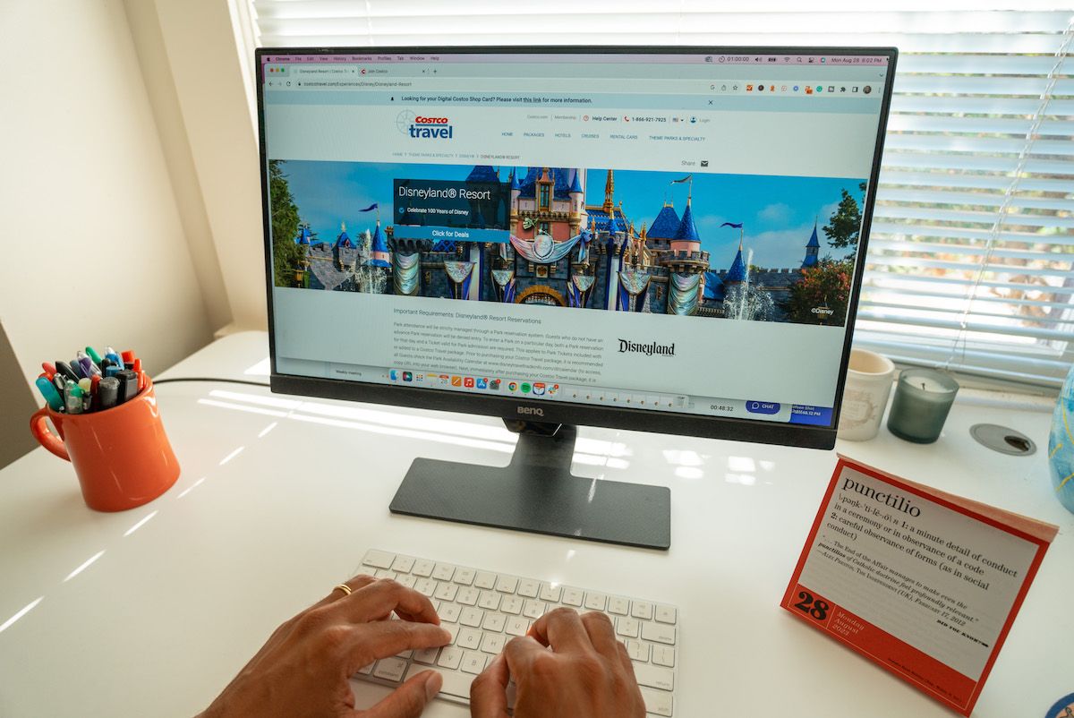 A pair of hands type on a keyboard sitting on a white desk in front of a monitor displaying a Costco Travel website page about Disneyland Resort.