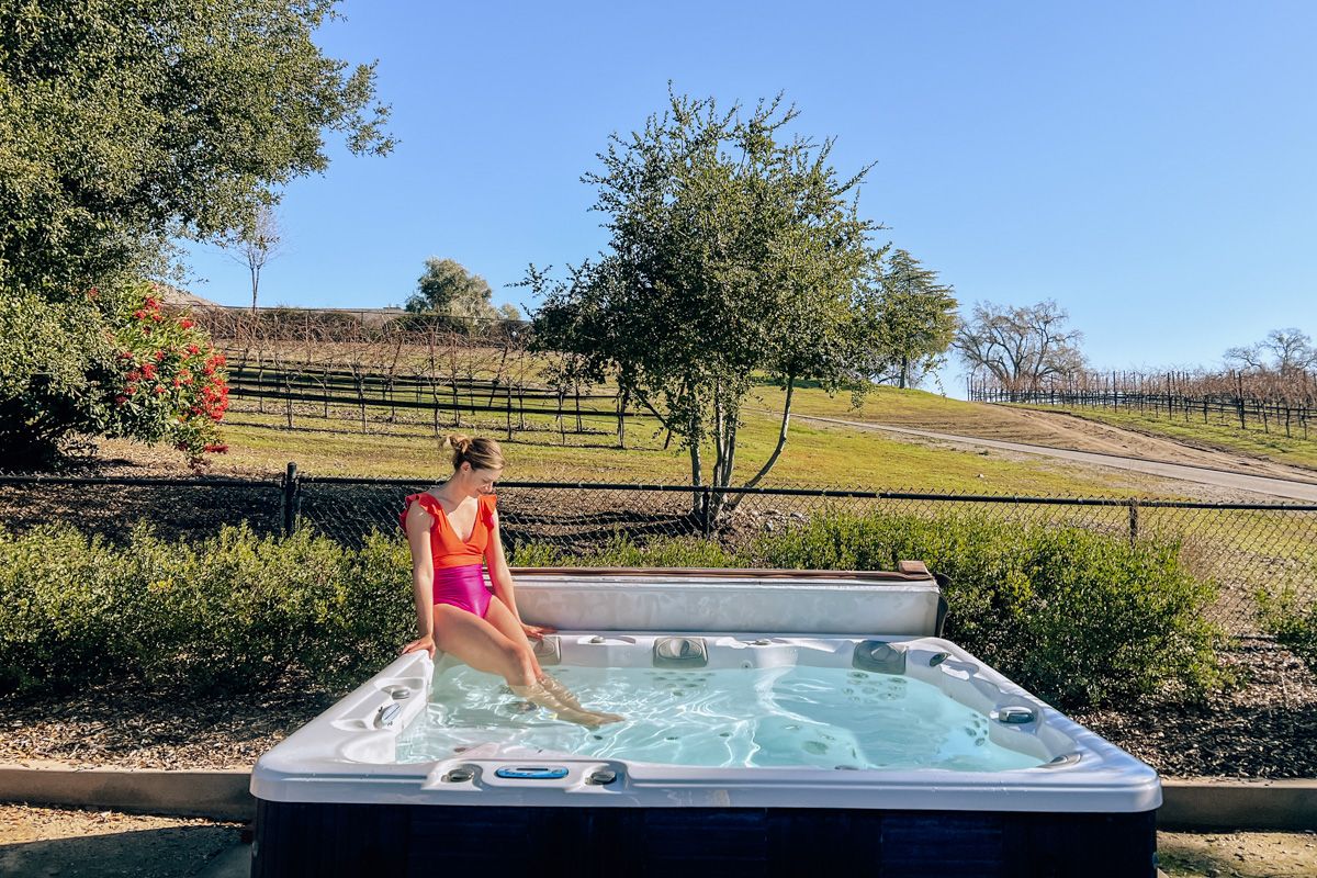 A young woman wearing an orange and magenta one-piece swimsuit sits on the edge of a hot tub with her feet in the water on a sunny day, with a chainlink fence and a small vineyard in the background.