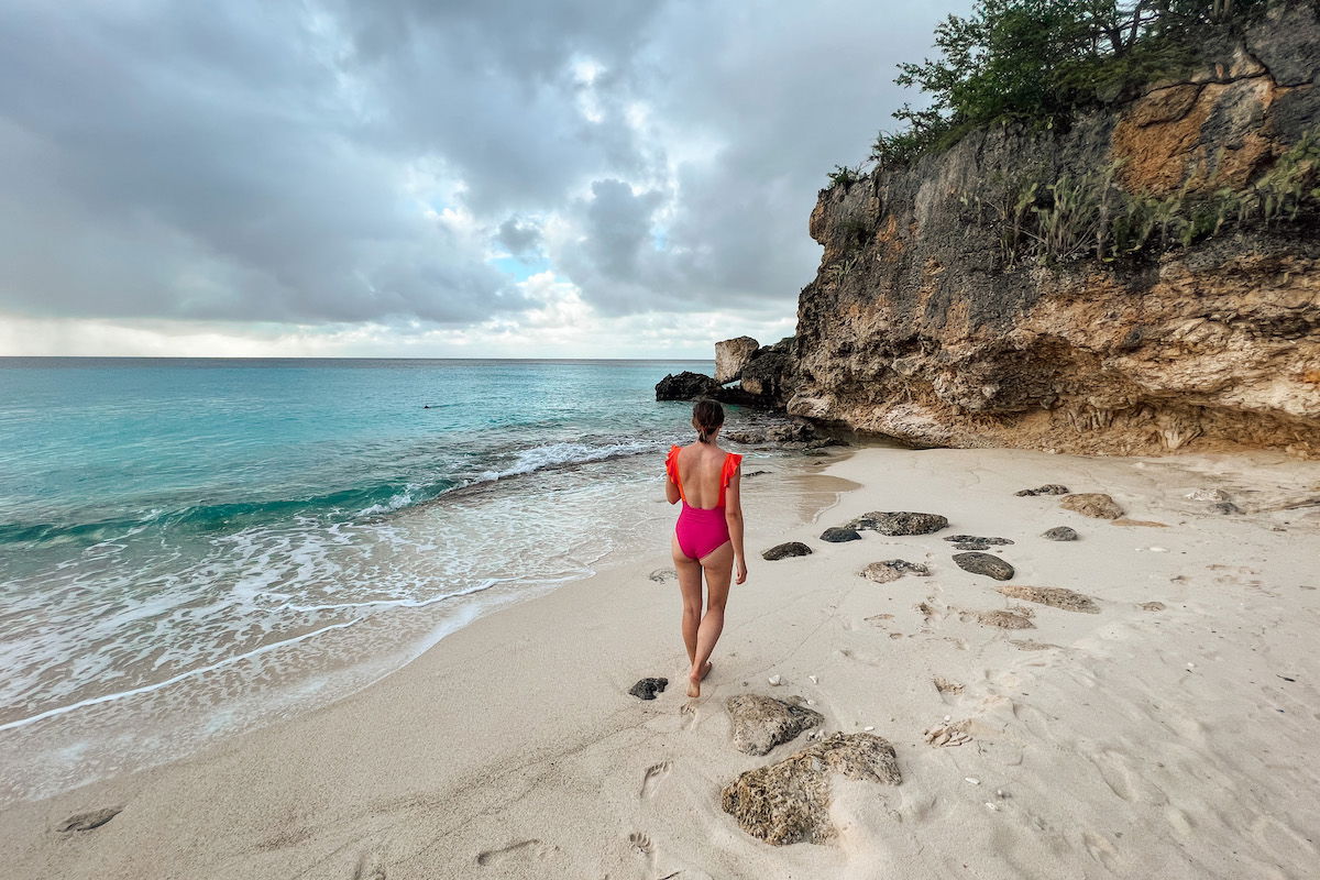 A young woman wearing an orange and magenta one-piece swimsuit seen from behind walking on a tropical-looking beach under an overcast sky.