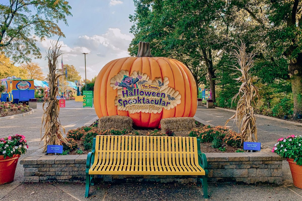 A yellow park bench in front of a large model of a pumpkin decorated with text that reads, "Count's Halloween Spooktacular At Sesame Place."