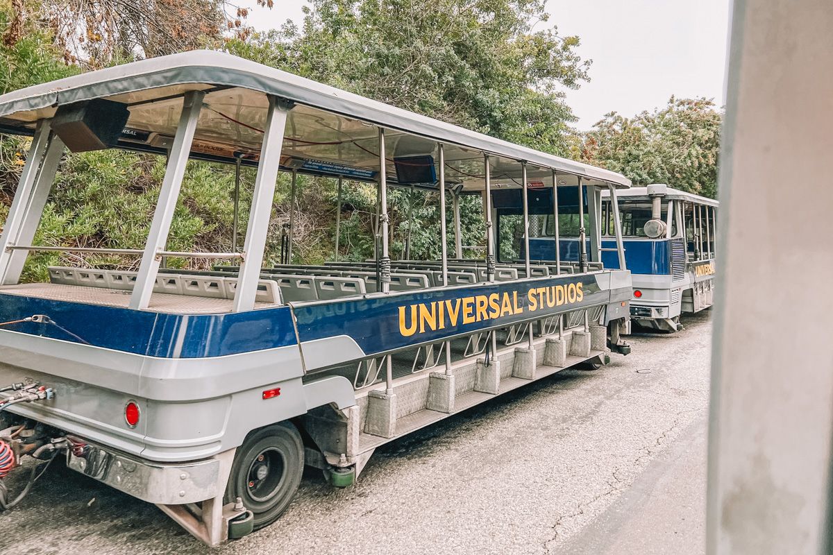 An open-side tour bus with the words, "Universal Studios" printed in yellow text against dark blue, parked by the side of the road with bushes behind it.