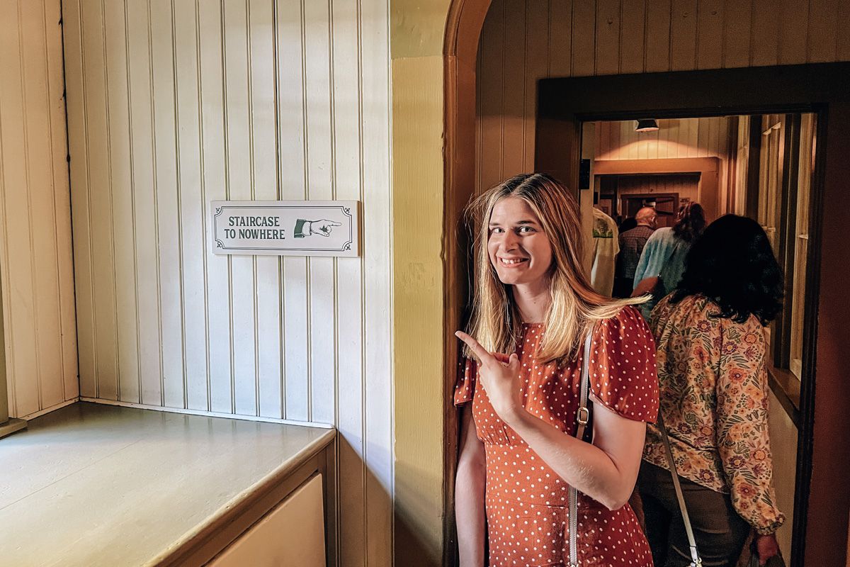 A young woman in a red polka-dot dress standing in a doorway inside the Winchester Mystery House and pointing at a sign reading "staircase to nowhere."