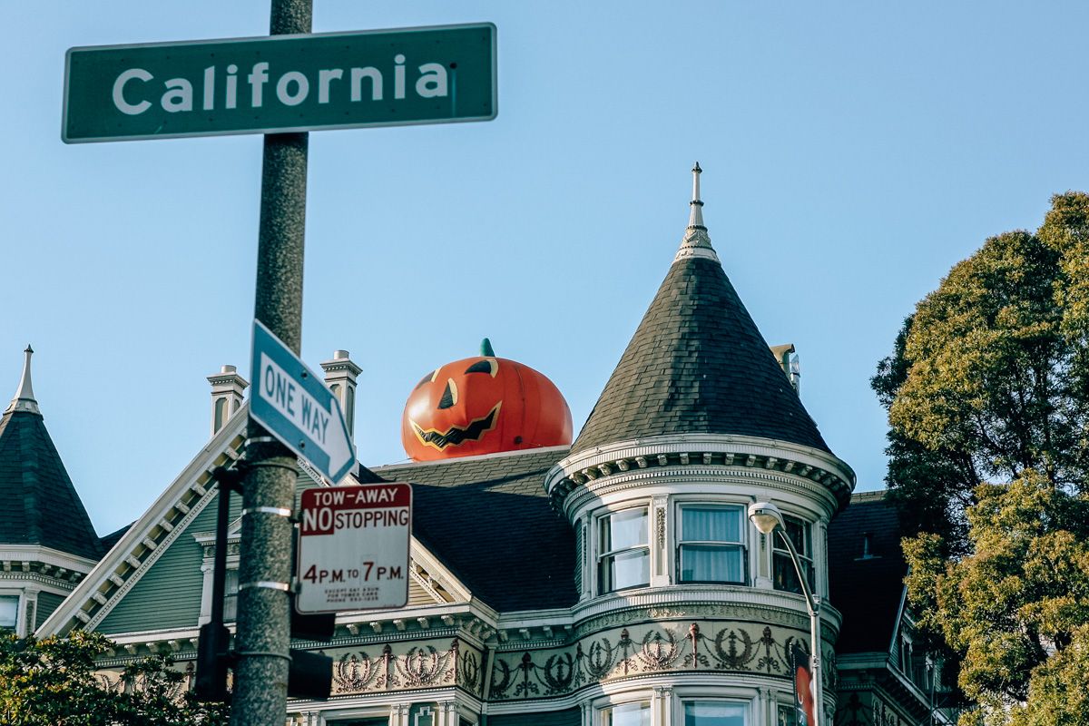 A San Francisco victorian house with a giant jack-o-lantern decoration perched on its roof, with a green street sign in the foreground that reads, "California."
