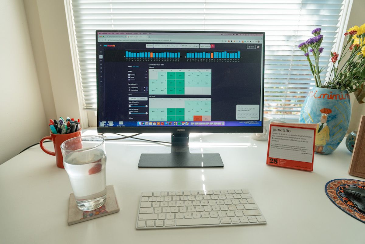 A view of a white desk with a computer monitor sitting on it, displaying the flight search results on the Momondo website, with a glass. ofwater sitting on a coaster beside it.