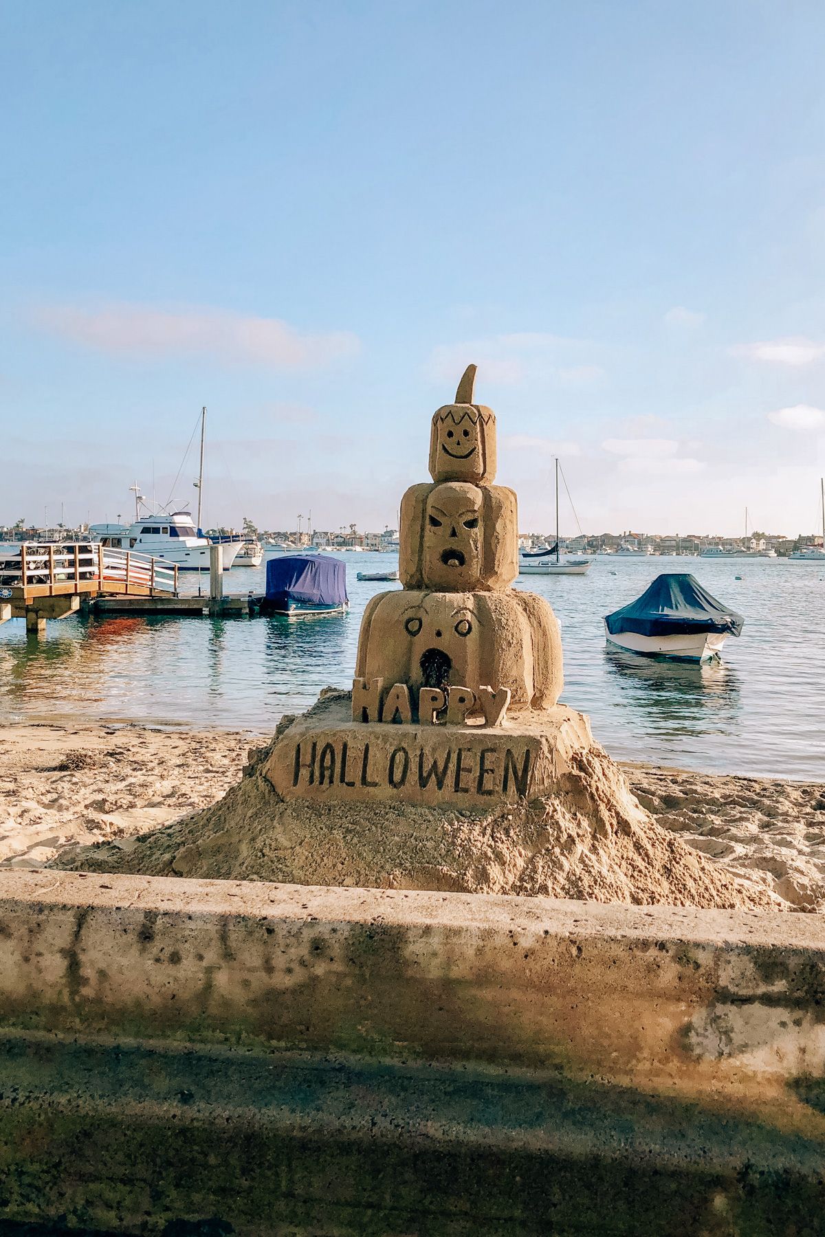 Halloween Events in Orange County: A Hallloween-themed sandcastle with the words, "Happy Halloween" carved into it on a beach with a a dock and boats in the background.