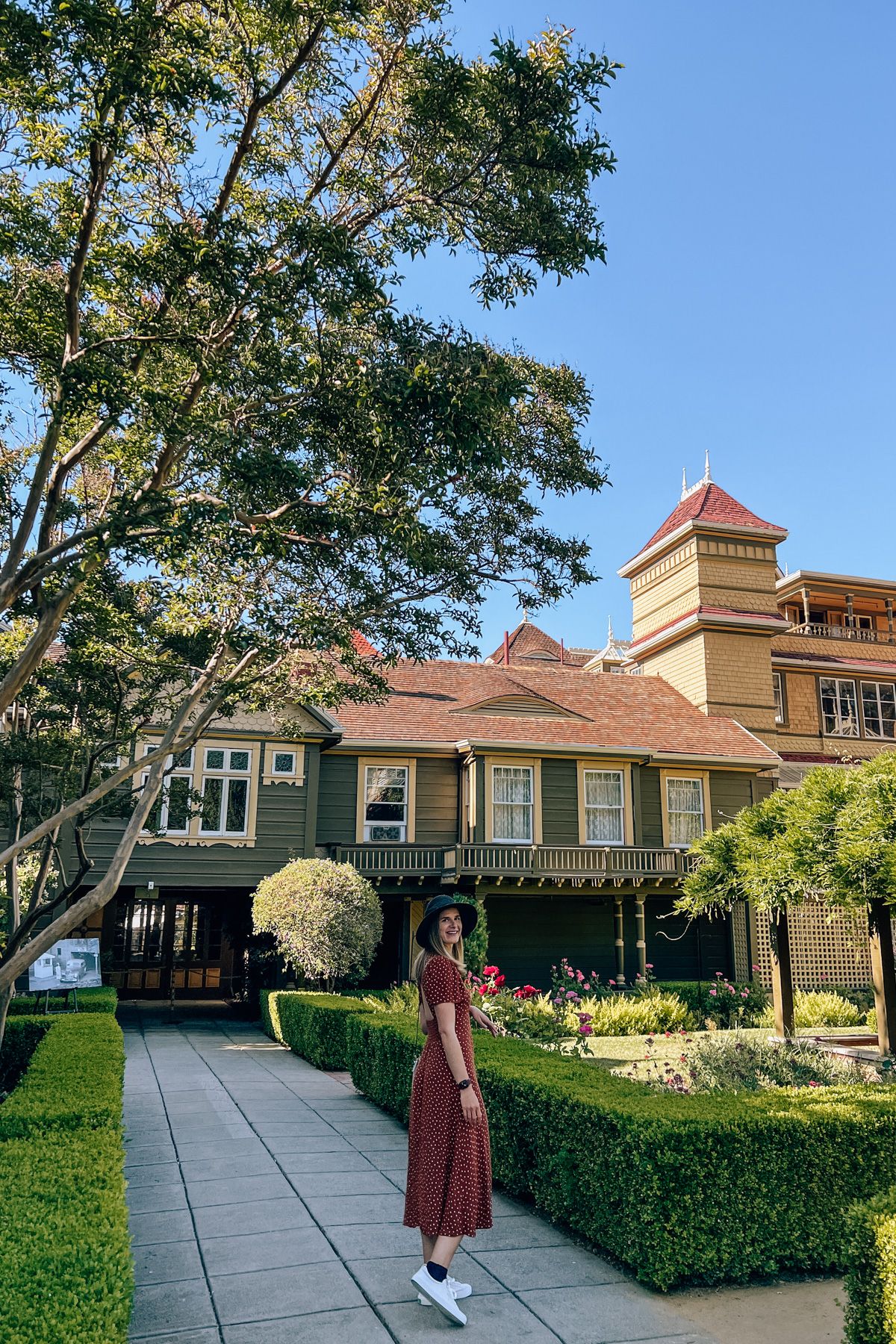 San Francisco Haunted Houses: A young woman in a red polka-dot dress and a wide-brimmed black hat looks back over her shoulder in the shaded, manicured garden of a victorian house.