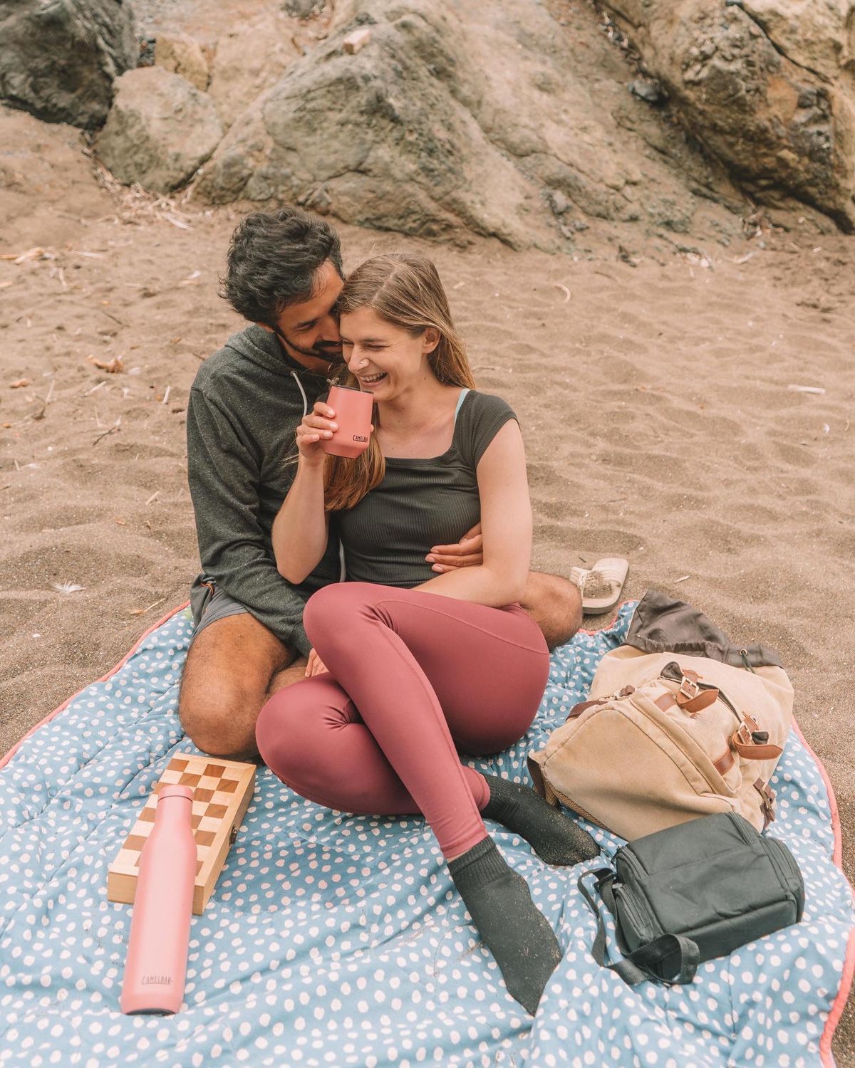 A young couple cuddles and laughs as they sit on a blue and white polka-dotted blanket on the sand, with several bags, a travel chessboard, and a water bottle spread out around them. 