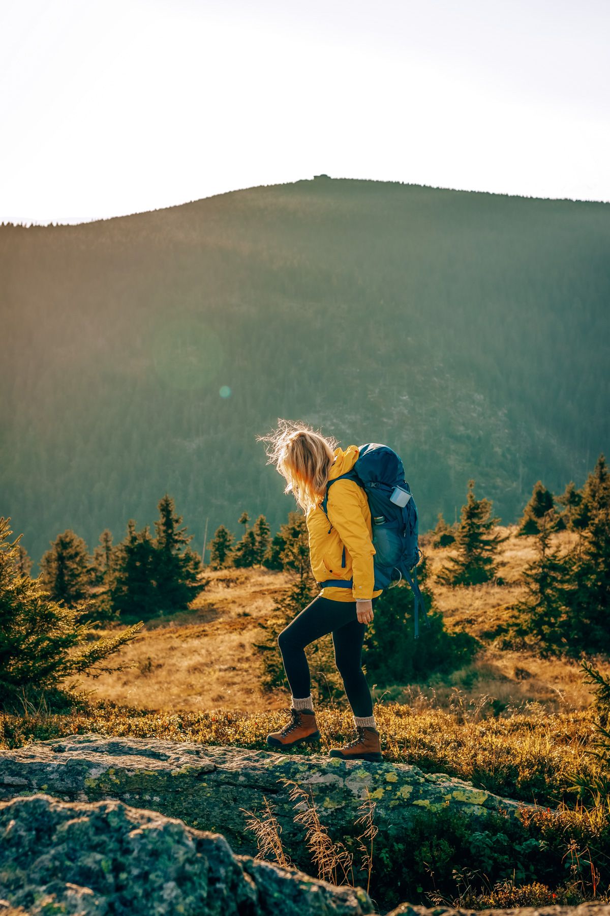 A blonde woman seen in profile wearing a yellow windbreaker, black leggings, and a blue backpack hiking over mountainous terrain in golden afternoon light.