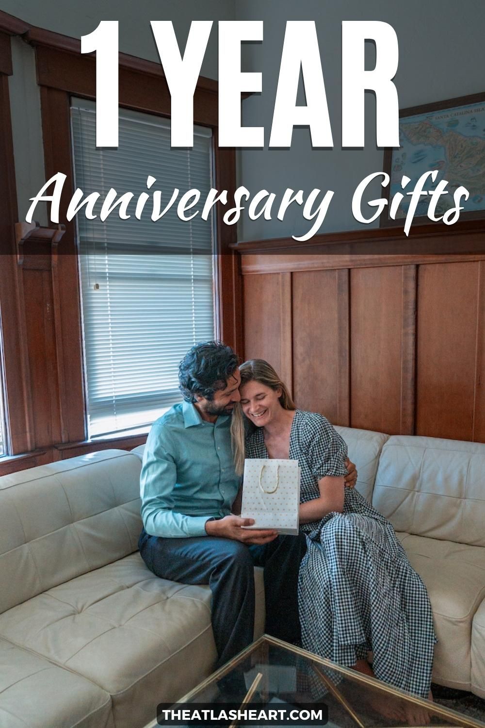 A man in a blue shirt embraces a woman in a gingham maxi dress and hands her a white gift bag  as they sit in the corner of an L-shaped, white leather couch, with a wood-paneled interior in the background and the text overlay, "1 Year Anniversary Gifts."
