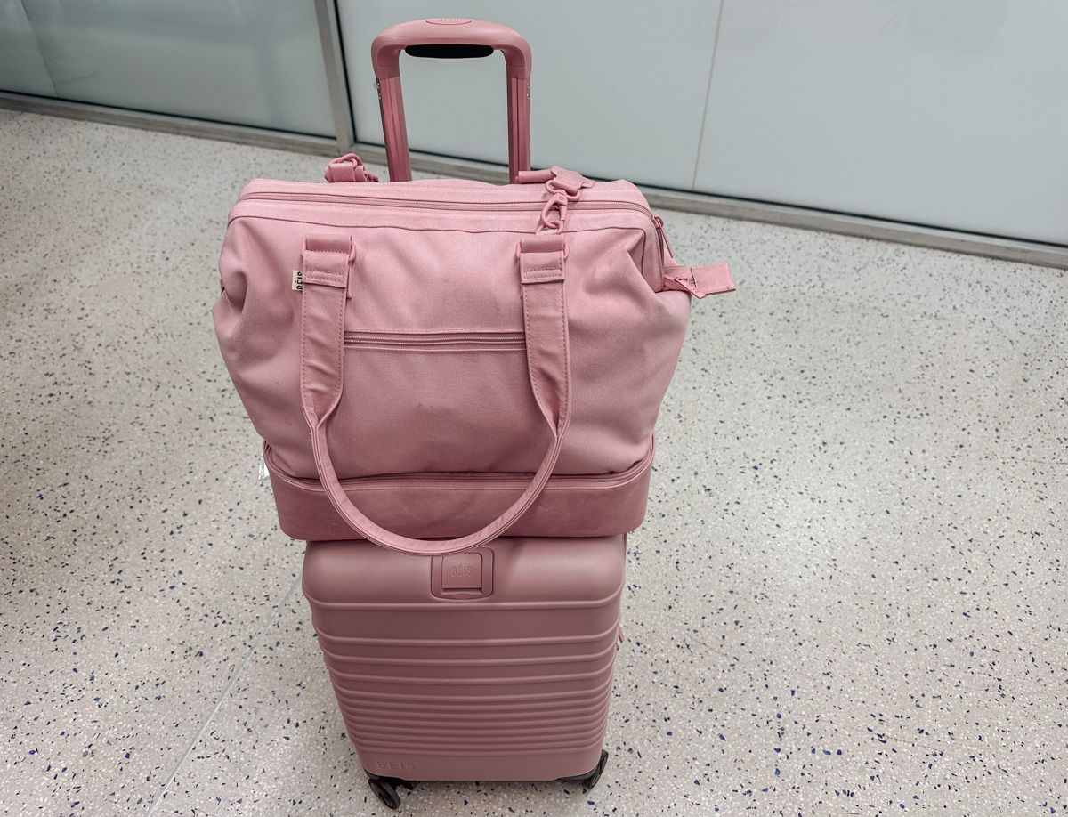 A pink Beis Mini Weekender sitting on top of a pink 21" Béis Luggage Carry On sitting on a grey-flecked airport floor.