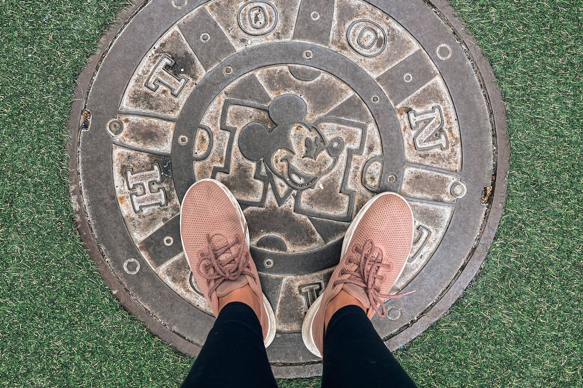 A POV-shot looking down at a pair of feet wearing pink Allbirds and standing on a Mickey Mouse manhole cover surrounded by astroturf.