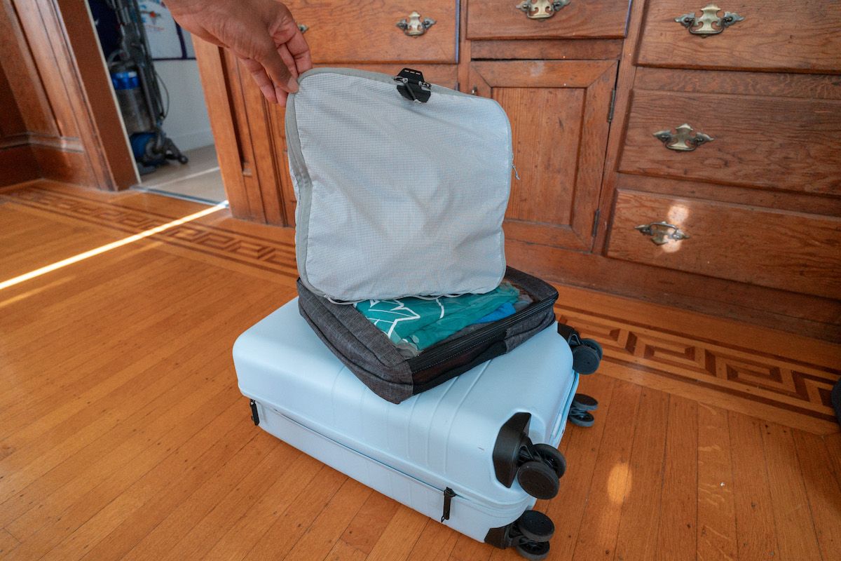 A light blue suitcase sits on a wooden floor with two packing cubes stacked on top of it, as a hand comes in from the top of the frame to hold one of them up.