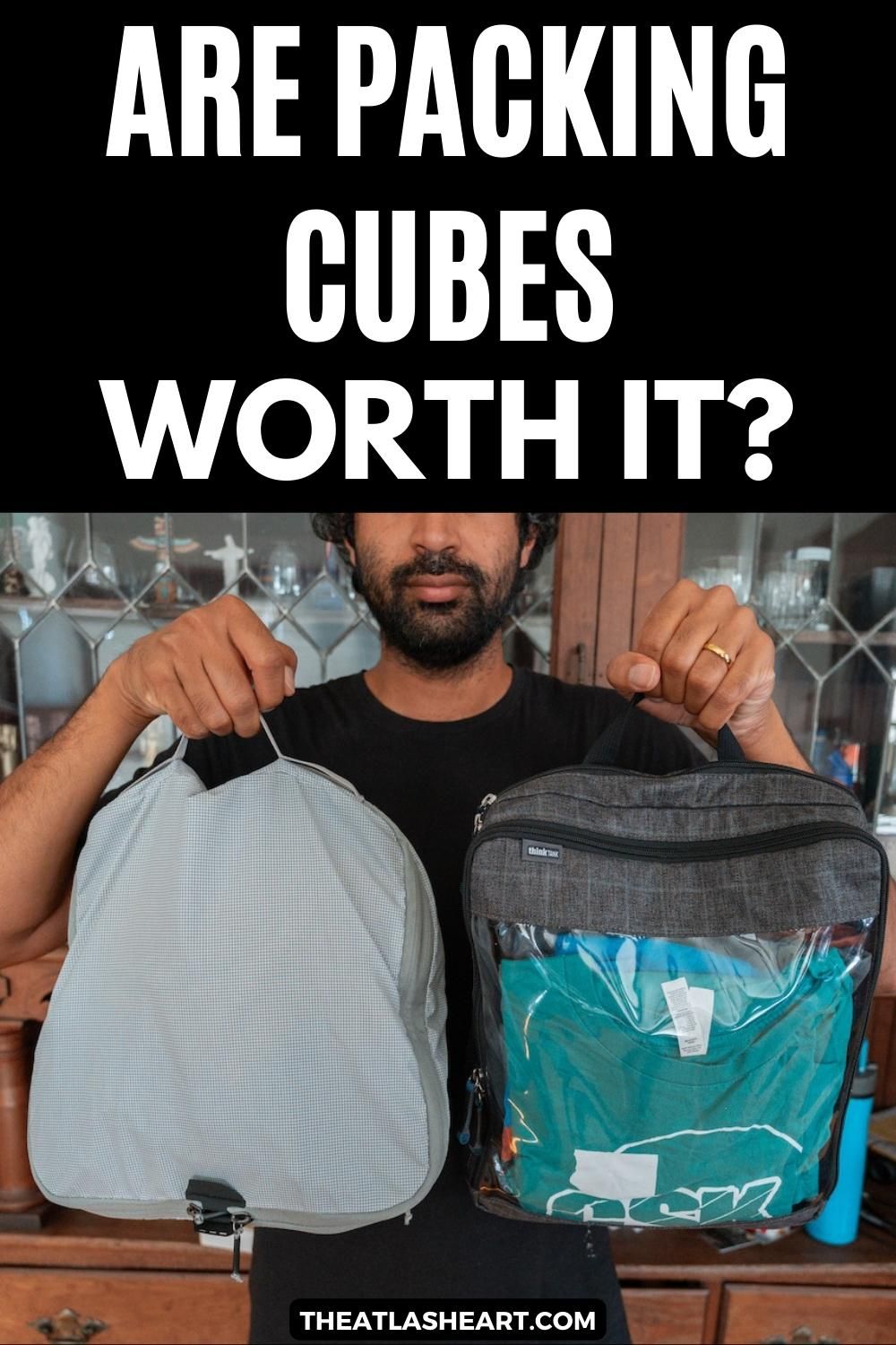 A picture of a man taken at chest level as he holds up two different styles of packing cubes with a wooden cabinet behind him, with the text overlay, "Are Packing Cubes Worth It?"