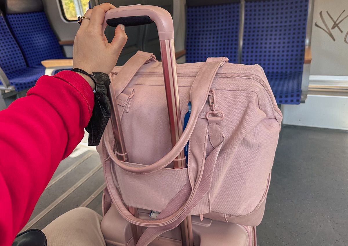 A POV-shot of an arm with a red sleeve holding the handle of a pink Beis Suitcase with a pink Beis Mini weekender bag sitting on top and the blue seats of a train interior in the background. 