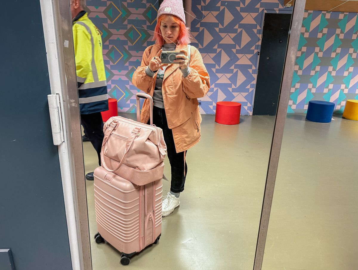 A young woman with pink hair, wearing a beanie and a salmon-colored coat takes a selfie in a train station mirror, with a pink Beis luggage set beside her and pink and purple patterned wallpaper in the background. 