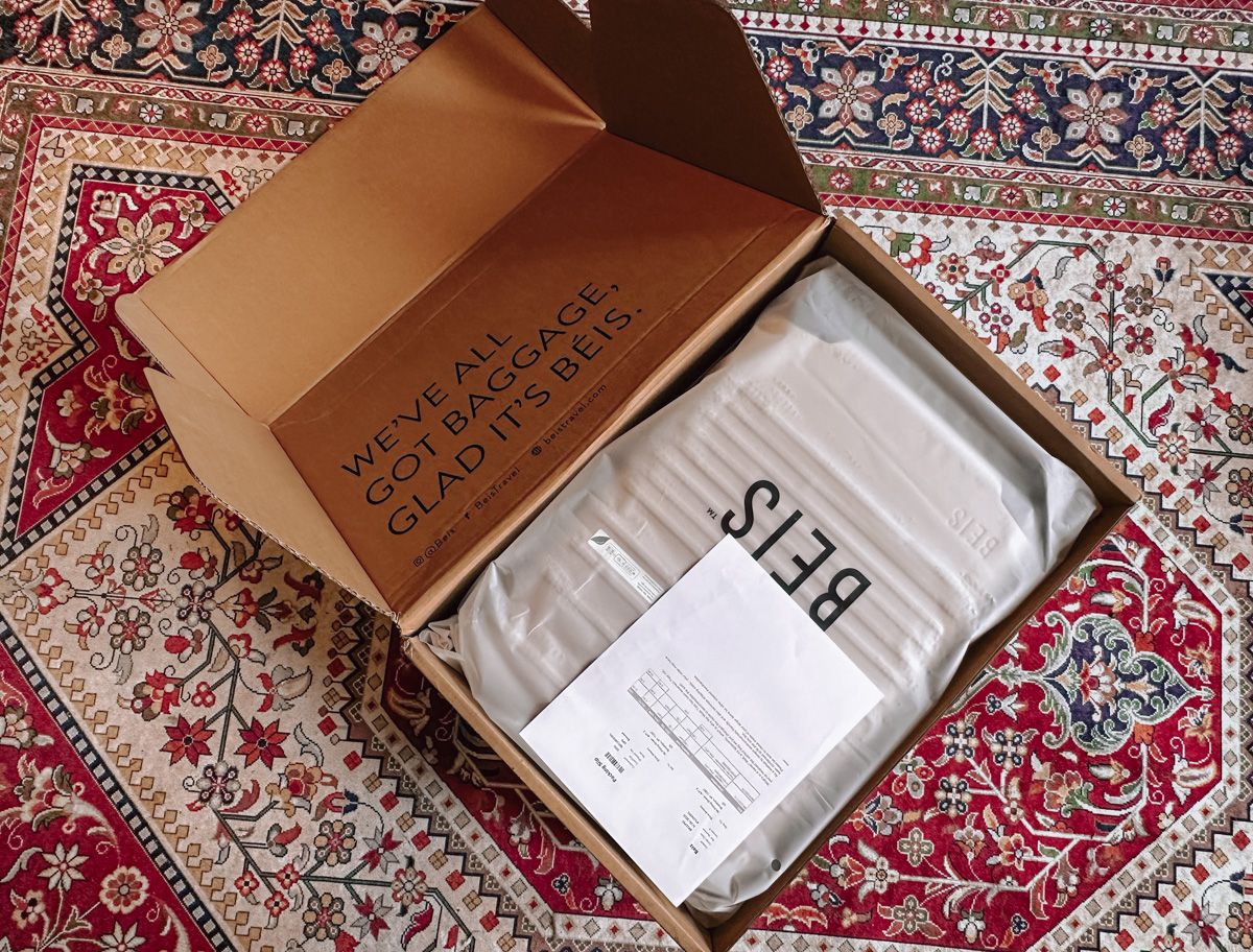 A carboard box sitting on a red, blue, and white oriental rug, open to reveal a packaged Beis suitcase inside.