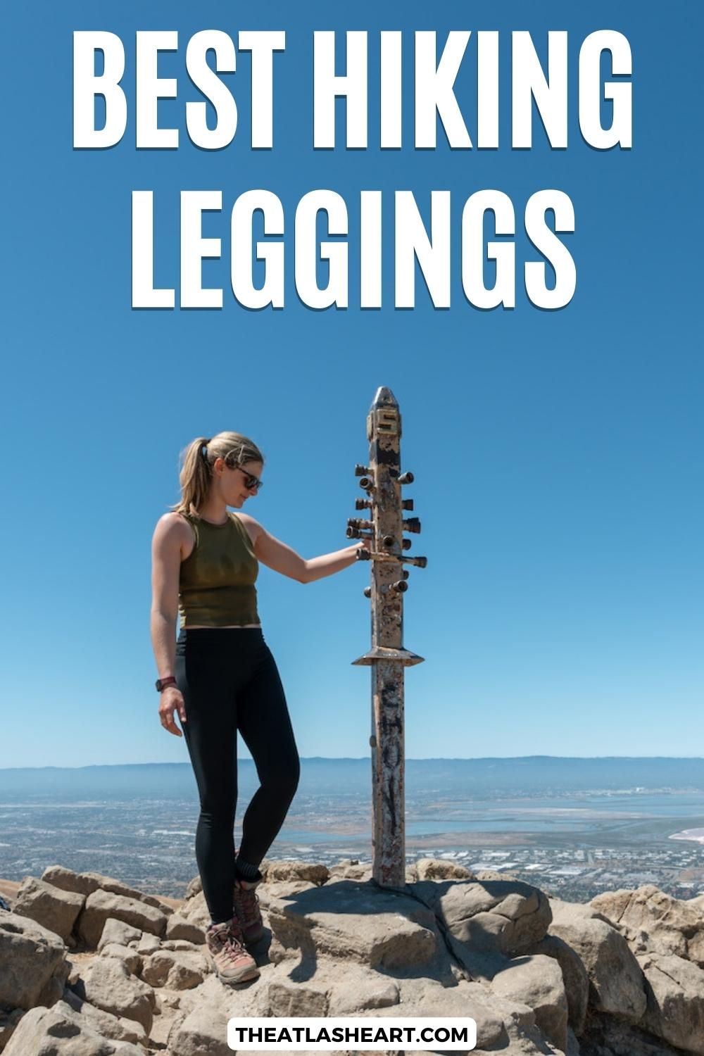 A woman in black leggings, a green tank, and hiking boots stands next to a totem-like pole at the rocky summit of a mountain overlooking a suburban valley, with  clear blue sky behind her, with the text overlay, "Best Hiking Legging."
