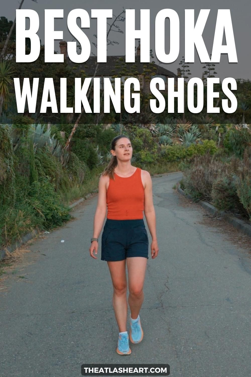 A young woman wearing light blue Hoka walking shoes, an orange tank top, and navy shorts walks towards the camera on a paved path lined by succulents and bushes, with the text overlay, "Best Hoka Walking Shoe."