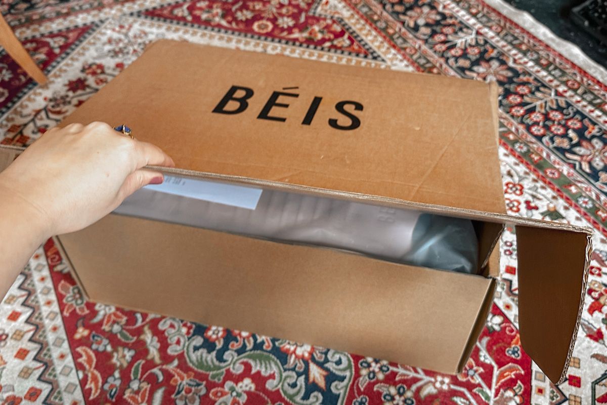 A hand opening a carboard box that says "Beis," sitting a red, blue, and white oriental rug. 