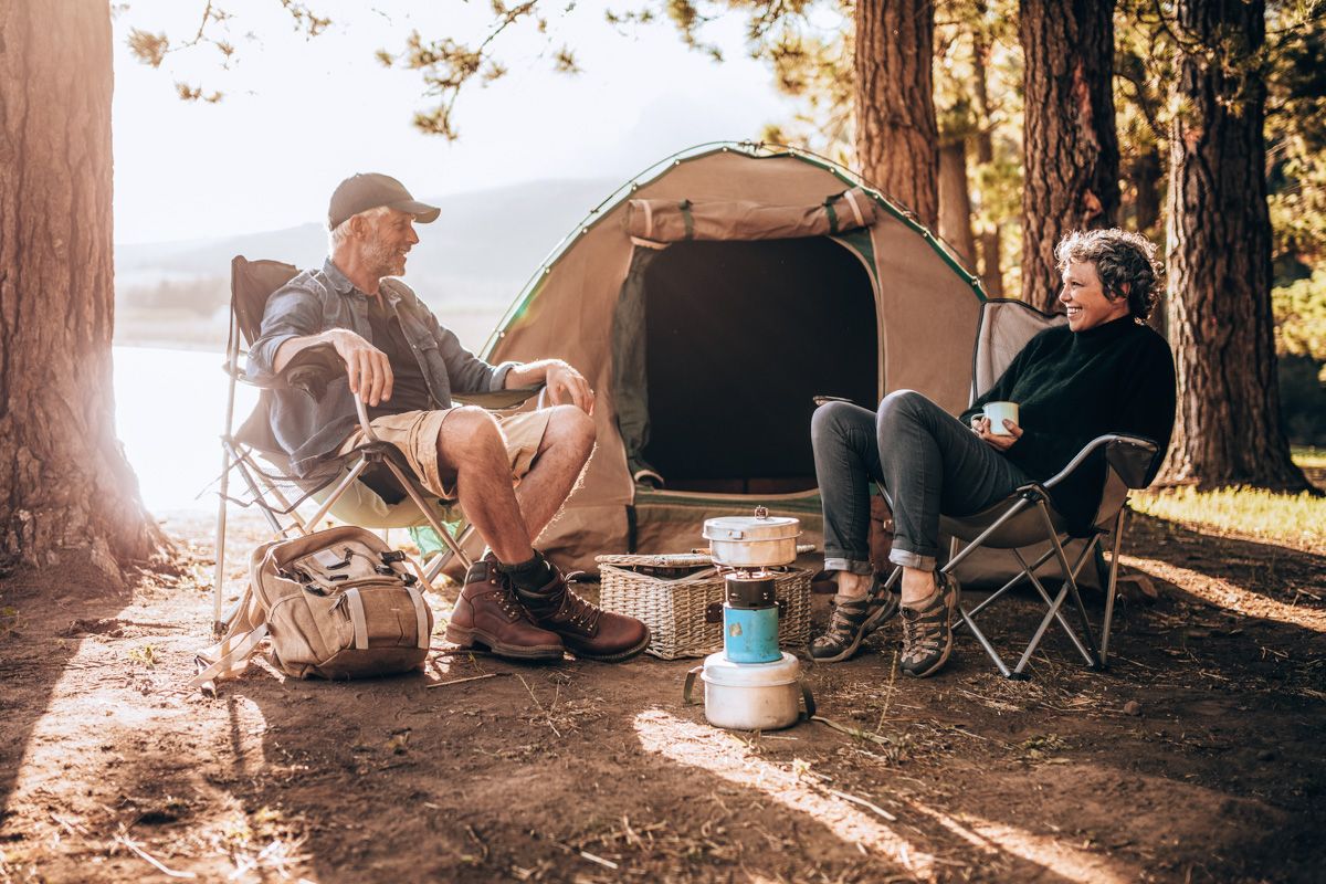 An older couple sitting in camping chairs smile at each other in a pine-tee studded campsite, with a darkroom tent visible behind them. 